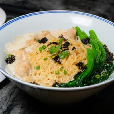 A bowl of Chinese noodles with dumplings, vegetables, and flavorful garnishes served indoors.